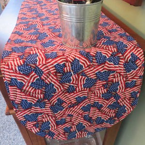 May include: A red, white, and blue table runner with a pattern of American flags. The runner is made of fabric and has a rounded end. A silver metal container is sitting on top of the runner.