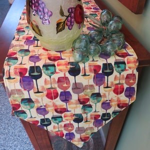 May include: A beige table runner with a repeating pattern of colorful wine glasses. The runner is decorated with a glass vase filled with grapes and a glass grape cluster.