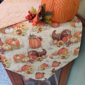 May include: A beige table runner with a fall theme featuring pumpkins, sunflowers, and cornucopias. The runner has a handwritten script pattern and is accented with red berries.