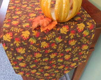May include: A brown table runner with a fall leaf pattern. The runner is decorated with orange, yellow, and red leaves. A yellow ceramic pumpkin sits on top of the runner.