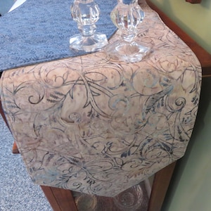 May include: A decorative table runner with a beige and blue floral pattern. The runner is draped over a wooden table, with two clear glass candle holders on top. A blue patterned fabric is visible on the table.
