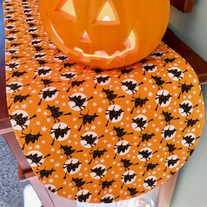 May include: Orange table runner with a repeating pattern of black witches flying on broomsticks in front of a white moon with white stars on an orange background.