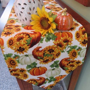 May include: A white table runner with a fall theme featuring pumpkins, sunflowers, and cornucopias. The runner is decorated with orange, yellow, and green colors.
