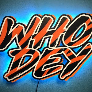 May include: A wall-mounted sign featuring the text WHO DEY in bold, stylized lettering. The letters have a black and orange tiger-stripe pattern with a white outline. A bright blue LED light glows from behind the sign, creating a halo effect.