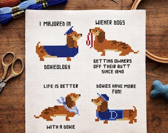 May include: A white cross-stitch fabric featuring four dachshund illustrations with text. The top left shows a dog in a graduation cap and gown with "I MAJORED IN DOXIEOLOGY". The top right shows a dog with a leash and "WIENER DOGS GETTING OWNERS OFF THEIR BUTT SINCE 1840". The bottom left shows a dog with a blue bandana and "LIFE IS BETTER WITH A DOXIE". The bottom right shows a dog in a blue shirt with "DOWIES HAVE MORE FUN!".