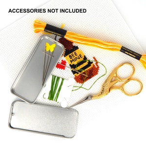 May include: A cross-stitch project on white Aida cloth featuring a bee design with the text BEE MINE, accompanied by yellow embroidery floss, gold-toned stork scissors, a green thread bobbin, two metal tins, and needles with a yellow butterfly topper.