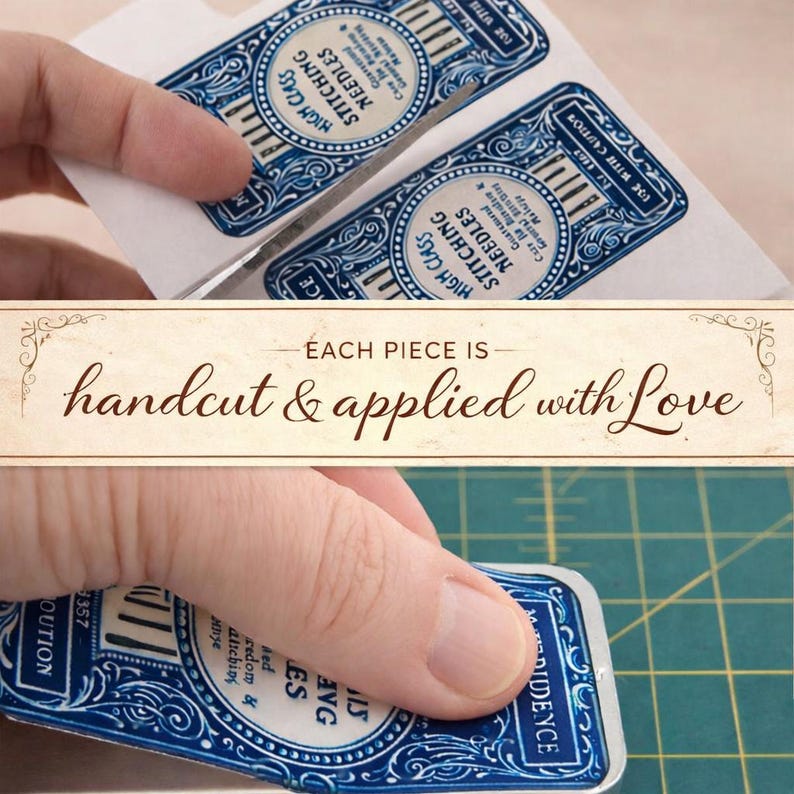 May include: A rectangular metal tin featuring a vintage-style blue and white label that reads High Class Stitching Needles. The label includes ornate decorative borders and text. A hand is shown applying the label to the surface of the metal container.