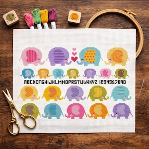 May include: A white cross-stitch fabric featuring rows of colorful elephants in pink, purple, blue, yellow, and green, with three small red hearts, the alphabet, and numbers 1-9 stitched in black, accompanied by embroidery floss and a hoop.