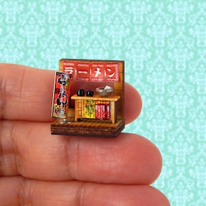 May include: Miniature wooden ramen shop with a red and white sign in Japanese. The shop features a small table with bowls and a menu board. The shop is brown with red, yellow, and black accents.