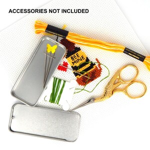 May include: A cross-stitch project on white Aida cloth featuring a bee design with the text BEE MINE, accompanied by yellow embroidery floss, gold-toned stork scissors, a green thread bobbin, two metal tins, and needles with a yellow butterfly topper.