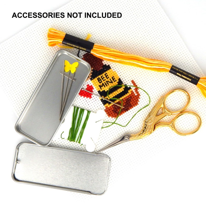 May include: A cross-stitch project on white Aida cloth featuring a bee design with the text BEE MINE, accompanied by yellow embroidery floss, gold-toned stork scissors, a green thread bobbin, two metal tins, and needles with a yellow butterfly topper.