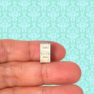 May include: A miniature, two-door oven, likely for a dollhouse or craft project. The oven is made of light-colored wood with a silver top and door handles. The oven is held between two fingers.