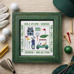 May include: A framed cross-stitch embroidery featuring golf-themed icons including a golf cart, clubs, and flags. The border text reads HOLE IN ONE, BIRDIE, PAR, EAGLE, and MULLIGAN. The piece is surrounded by white golf balls, tees, and a golf club.