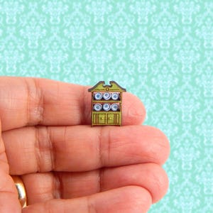 May include: Miniature wooden cabinet painted in shades of green and brown, with a decorative top and two doors. The cabinet has shelves holding small, round plates with blue and white designs. A charming dollhouse accessory.