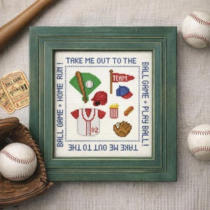 May include: A framed cross-stitch embroidery featuring baseball-themed icons including a bat, ball, helmet, jersey, pennant, popcorn, hot dog, and glove. The text reads "TAKE ME OUT TO THE BALL GAME" and "PLAY BALL!" in blue, surrounded by a green frame.