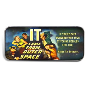 May include: A silver metal pin with a colorful image of a movie poster. The poster features the text "IT CAME FROM OUTER SPACE" and a group of people looking scared. The text below the image reads "If you've ever wondered why your stitching needles feel odd. Maybe it's because..."