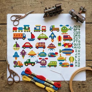 May include: A white cross-stitch fabric featuring a grid of colorful embroidered vehicles including trains, cars, planes, and boats, with a column of alphanumeric text on the right. The fabric is surrounded by metal scissors, embroidery floss, and a wooden hoop.