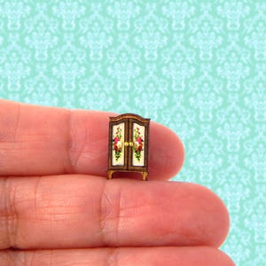 May include: A miniature wooden cabinet with two doors, painted with a floral design in shades of pink, white, and green. The cabinet has gold-colored knobs and sits on a wooden base.