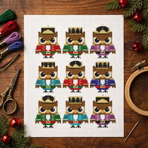 May include: A cross stitch pattern on white fabric featuring nine pixelated owls dressed as nutcrackers in various colors including red, green, purple, and blue. Each owl wears a unique hat and uniform, arranged in a three-by-three grid layout.
