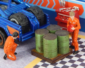 Puede incluir: Escena de garaje en miniatura con un camión azul, barriles verdes y dos figuras con monos naranjas. La escena incluye una caja de herramientas y un suelo a cuadros, creando un diorama detallado.