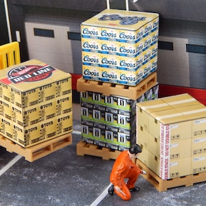 May include: Miniature boxes stacked on wooden pallets. One box features "RED LINE CLUB TOYS" text. Other boxes display "Coors" branding. A small figure in an orange jumpsuit kneels nearby. The scene suggests a warehouse or shipping theme.