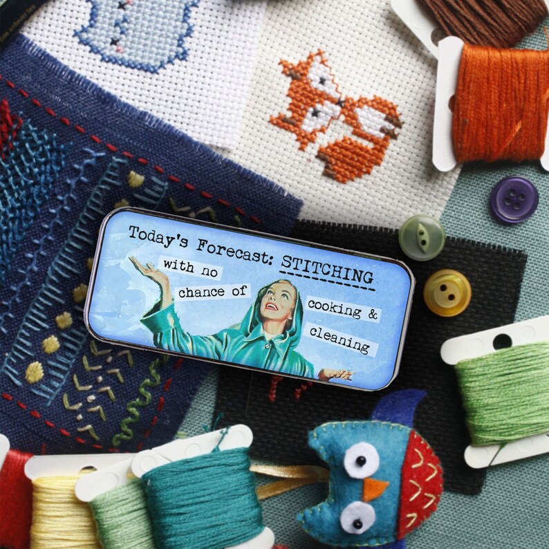 May include: A rectangular metal tin featuring a vintage-style illustration of a person in a teal raincoat looking upward. The text reads: Today's Forecast: STITCHING with no chance of cooking & cleaning. The tin is surrounded by embroidery supplies.
