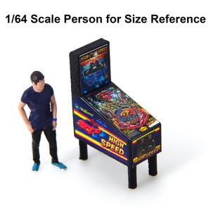 May include: Miniature pinball machine with a colorful graphic design, featuring the text "High Speed." The machine has black legs and a detailed playfield. A small figure is included for size reference.