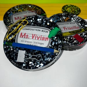 May include: Three round, silver glitter, composition book covers with teacher names and school supplies. The covers are labeled "Composition Book" with the teacher's name, number of sheets, and pages. The supplies include a pencil, crayons, and paper clips.