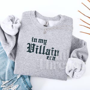 May include: A heather gray sweatshirt with the phrase "in my Villain era" printed in dark green gothic lettering. A silver necklace with a sunburst pendant rests on the shirt. The sleeves are cuffed, and the shirt is laid on top of a pair of blue jeans.
