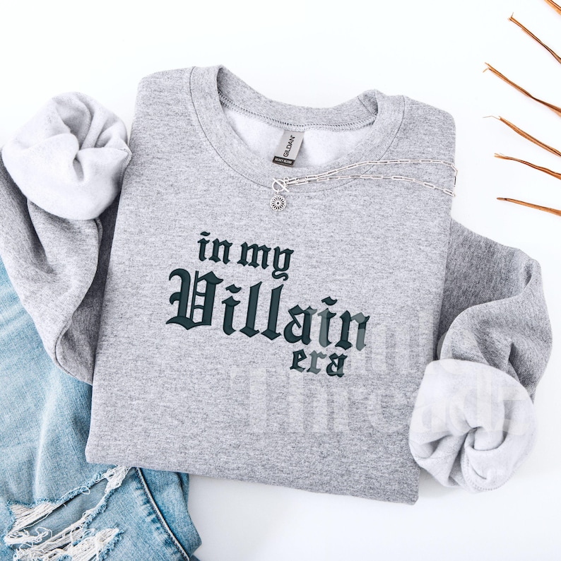 May include: A heather gray sweatshirt with the phrase "in my Villain era" printed in dark green gothic lettering. A silver necklace with a sunburst pendant rests on the shirt. The sleeves are cuffed, and the shirt is laid on top of a pair of blue jeans.