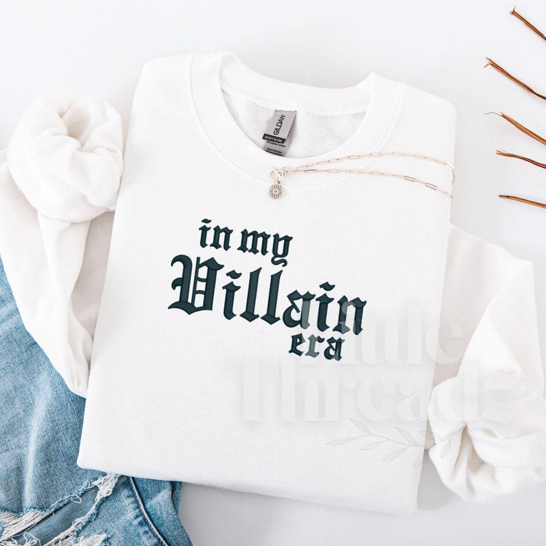 May include: White sweatshirt with the text "in my Villain era" in a dark teal gothic font. A silver necklace with a small pendant rests on the sweatshirt. The sweatshirt is folded on top of a pair of blue jeans.