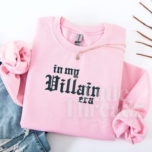 May include: A light pink sweatshirt with the phrase "in my Villain era" in dark green gothic lettering. A delicate necklace with a sunburst pendant rests on the sweatshirt. The sleeves are folded, and a pair of blue jeans are visible.