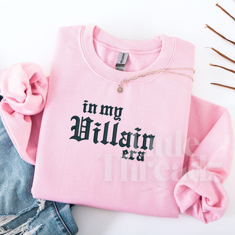May include: A light pink sweatshirt with the phrase "in my Villain era" in dark green gothic lettering. A delicate necklace with a sunburst pendant rests on the sweatshirt. The sleeves are folded, and a pair of blue jeans are visible.