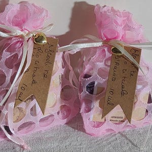 Può includere: Due sacchetti di rete rosa legati con nastri bianchi, ciascuno con un piccolo ciondolo a forma di cuore dorato. Le borse contengono piccoli oggetti e hanno etichette marroni con testo scritto a mano. Il testo sulle etichette recita "Ascooli re dio itaad 10 ml" e "Este to rituale D'Amora 10 ml".