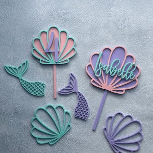 May include: A set of cake toppers and decorations featuring two shell-shaped toppers with sticks, one displaying the number 4 and the other the name Isabelle. Includes two mermaid tail cutouts and two additional shell cutouts in pastel colours.