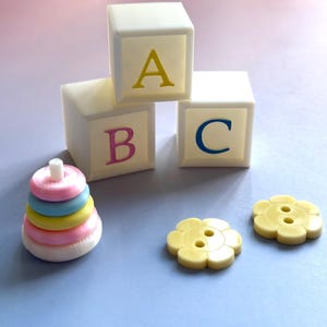 May include: A collection of children's toys on a blue surface. Three off-white alphabet blocks with the letters A, B, and C in yellow, pink, and blue. A stacking ring toy and two yellow flower-shaped buttons are also visible.