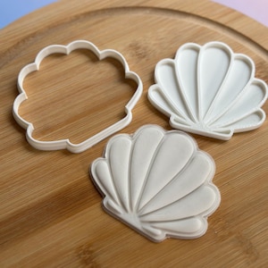 May include: Three white seashell-shaped cookie cutters on a wooden board. One is an outline, and the other two have detailed shell designs. The cutters are made of plastic and are suitable for baking themed biscuits.