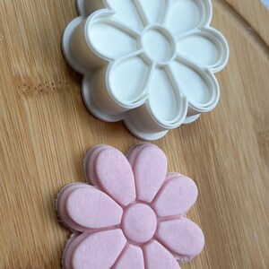 Pode incluir: Um cortador de biscoitos de flores de pl&aacute;stico branco e um biscoito de flores de fondant rosa. O cortador de biscoitos est&aacute; em cima do biscoito.