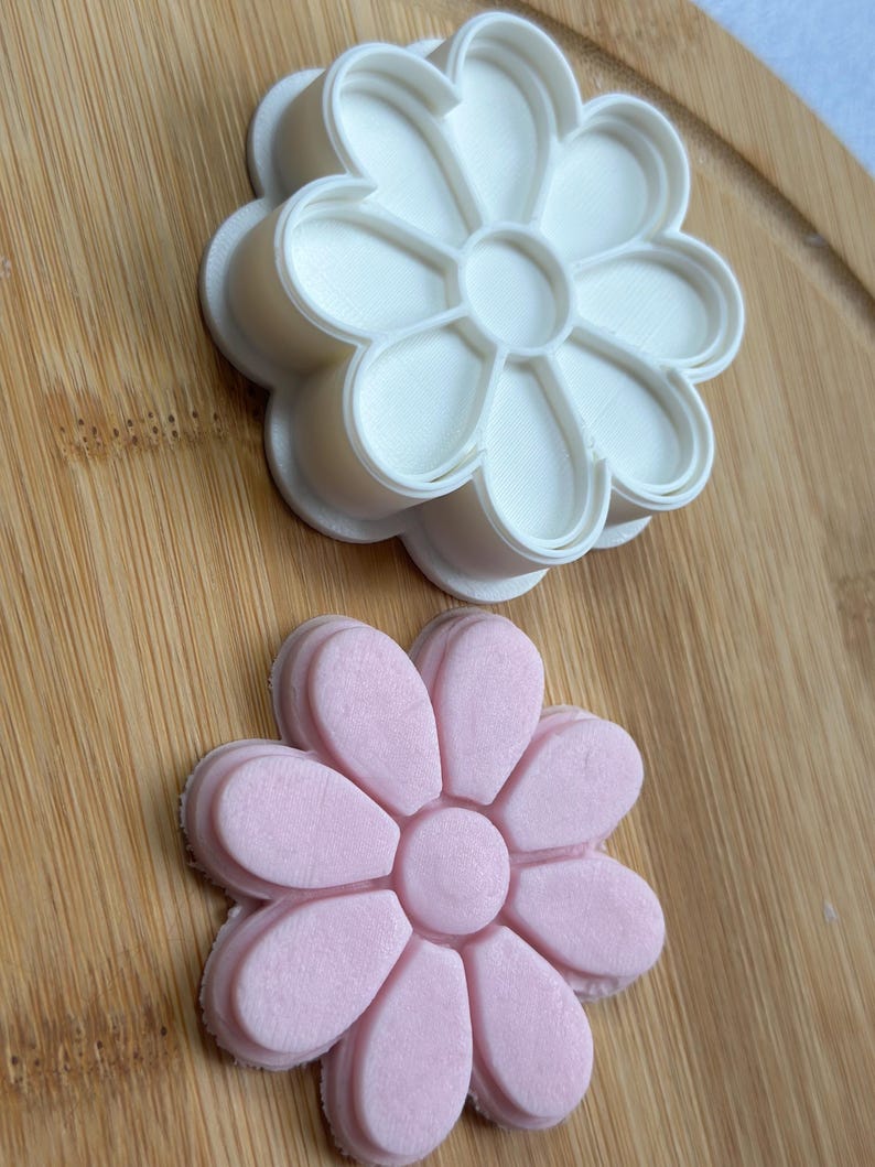 Pode incluir: Um cortador de biscoitos de flores de pl&aacute;stico branco e um biscoito de flores de fondant rosa. O cortador de biscoitos est&aacute; em cima do biscoito.