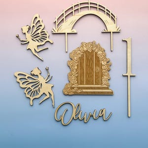 May include: Wooden fairy-themed cake toppers and decorations. Includes two fairy figures, a bridge, a door with floral accents, the name "Olivia", and the number "1". The items are made of light-coloured wood.