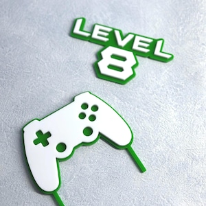 May include: White and green gaming-themed cake toppers. One topper reads "LEVEL 8" in white letters with a green outline. The other is a white controller with green accents and two green sticks.