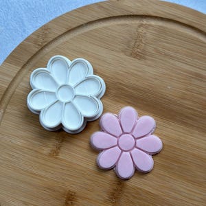 Pode incluir: Dois cortadores de biscoitos em forma de margarida, um branco e outro rosa, em uma t&aacute;bua de cortar de madeira.