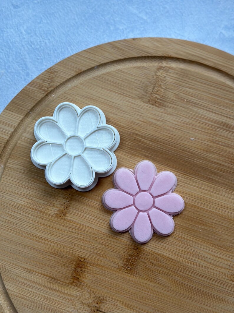 Pode incluir: Dois cortadores de biscoitos em forma de margarida, um branco e outro rosa, em uma t&aacute;bua de cortar de madeira.