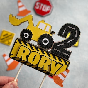 May include: A yellow construction vehicle cake topper with a black road number 2, a yellow and black construction sign with the name "Rory" and orange traffic cones.