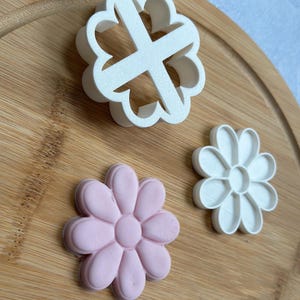 Pode incluir: Tr&ecirc;s cortadores de biscoitos em forma de flores sobre uma superf&iacute;cie de madeira. Um cortador &eacute; branco, outro &eacute; rosa e o &uacute;ltimo &eacute; branco com uma forma de cruz no centro.