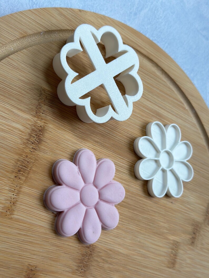 Pode incluir: Tr&ecirc;s cortadores de biscoitos em forma de flores sobre uma superf&iacute;cie de madeira. Um cortador &eacute; branco, outro &eacute; rosa e o &uacute;ltimo &eacute; branco com uma forma de cruz no centro.