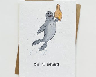 May include: A greeting card featuring a cartoon illustration of a seal giving a thumbs up with a foam finger. The text on the card reads "SEAL OF APPROVAL."