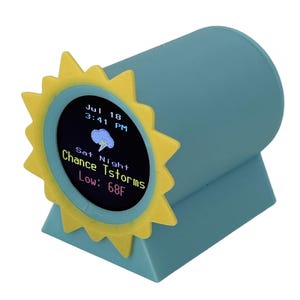 May include: A teal-colored weather station with a yellow sun-shaped frame. The digital display shows the date, time, and weather forecast: "Chance Tstorms, Low: 68F". The device has a cylindrical body and a triangular base.