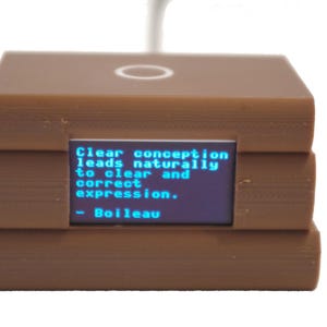 May include: A brown, rectangular device with a small screen displaying the quote "Clear conception leads naturally to clear and correct expression." The device has a circular indentation on top and a white background.