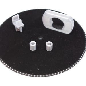 May include: A collection of small, silver-colored 3D-printed objects on a black circular surface. The objects include a small chair, a rectangular piece with a hole, and two cylindrical pieces. The edge of the circle has a white and black striped pattern.